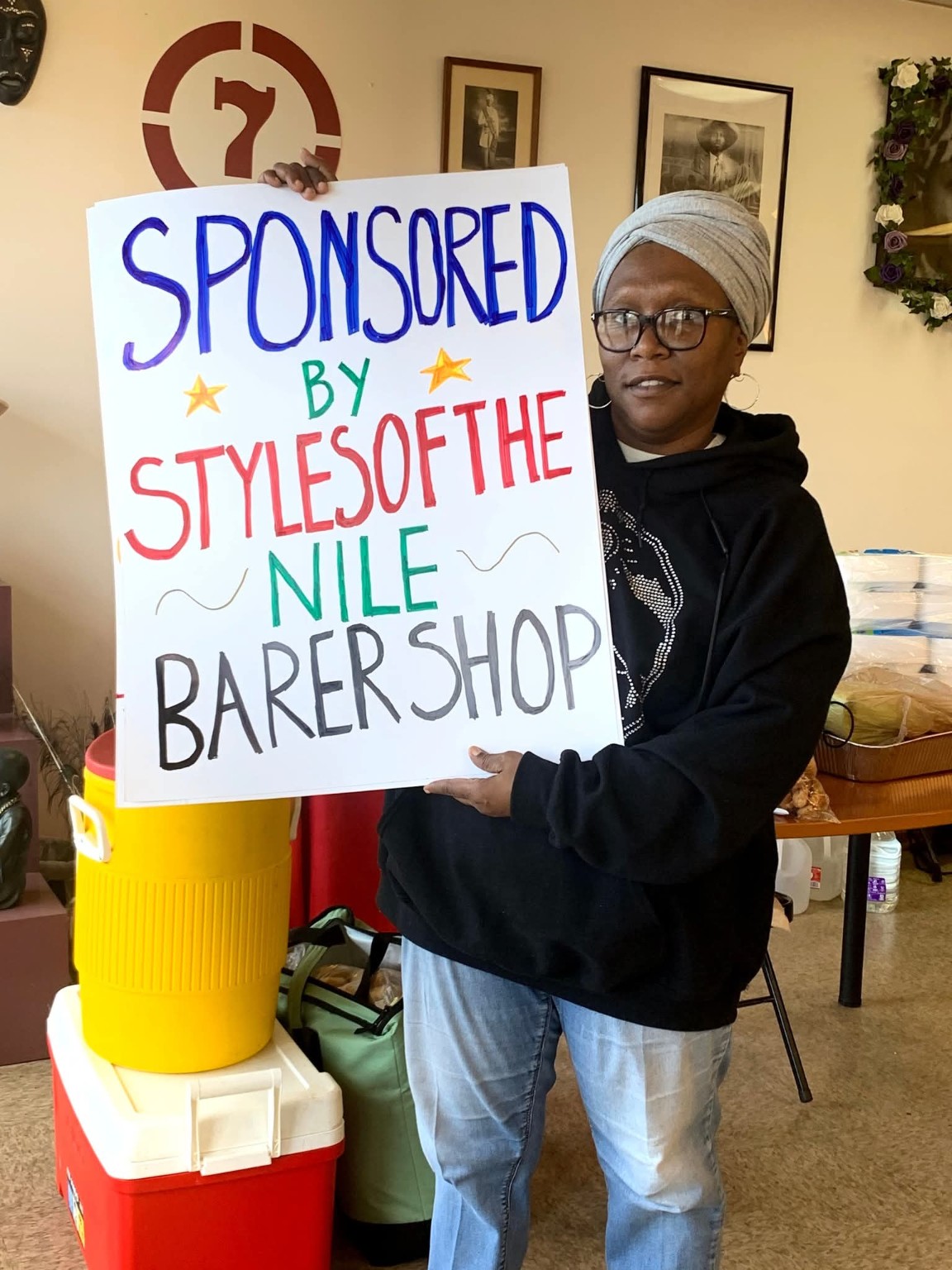 The barber’s wife holding a Sponsored by Styles of the Nile Barbershop sign